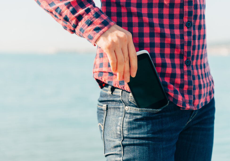 Woman takes out mobile phone of her pocket of jeans on beach near the sea to make self-portrait or to photograph the sea